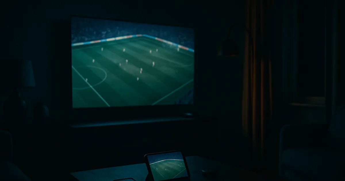 Mørkt dansk stue om aftenen oplyst af et stort tv, der viser en live fodboldkamp, mens samme kamp kører synkront på en telefon og en tablet på sofabordet.
