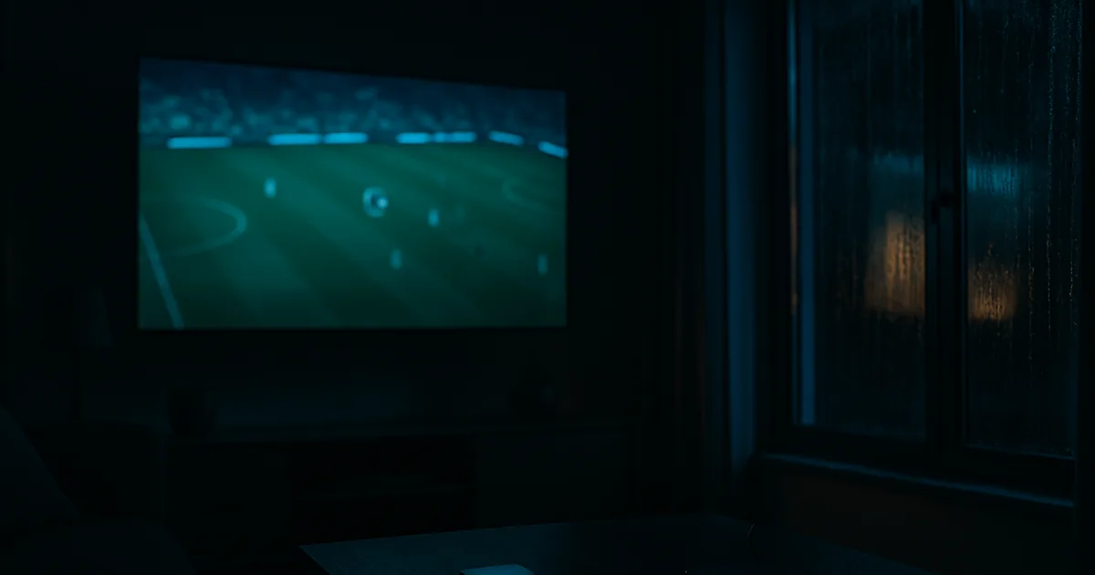 Mørk dansk stue om aftenen oplyst af et stort tv, der viser en fodboldkamp, med en streaming-boks og en telefon på sofabordet foran.